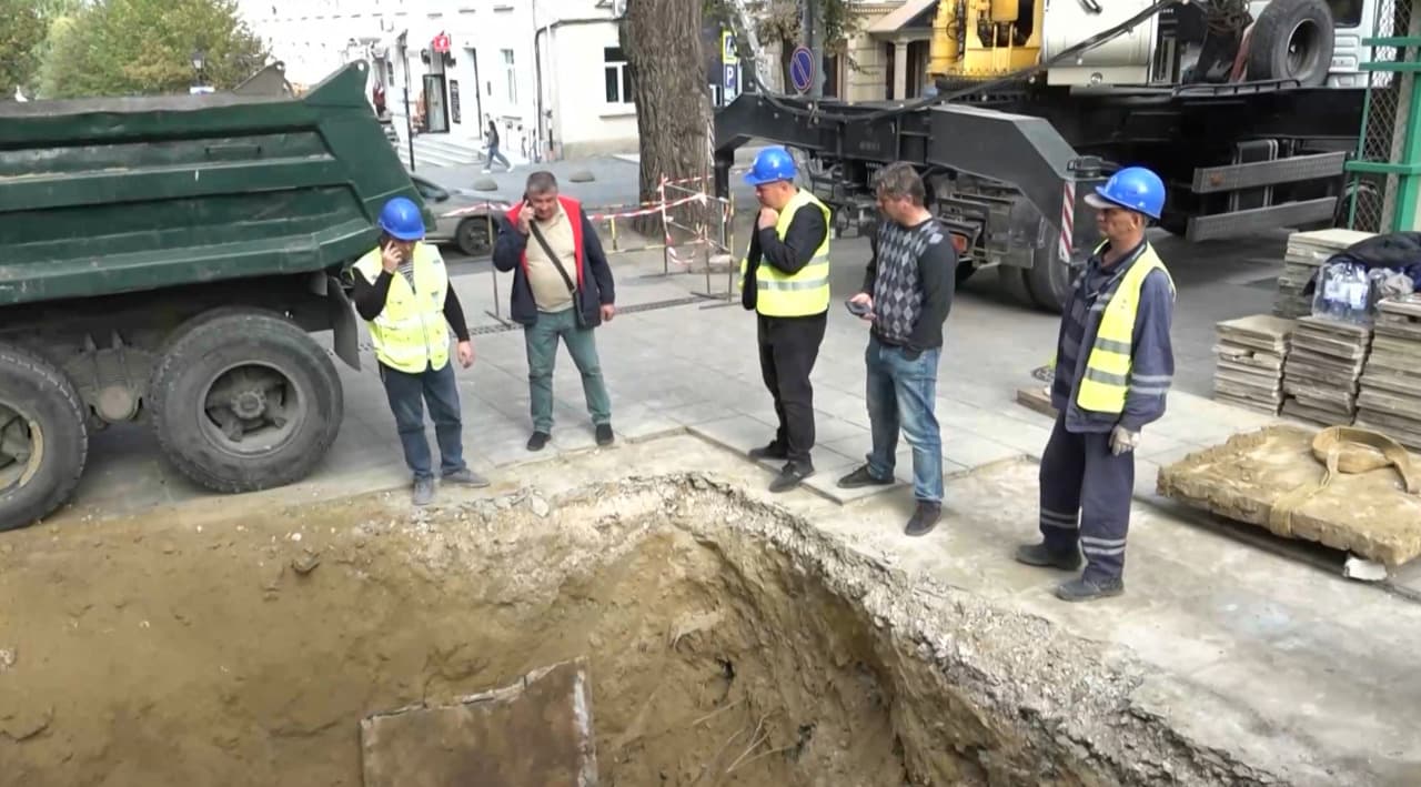 Muncitori în căști și veste reflectorizante lângă o groapă săpată în trotuar, lângă un camion